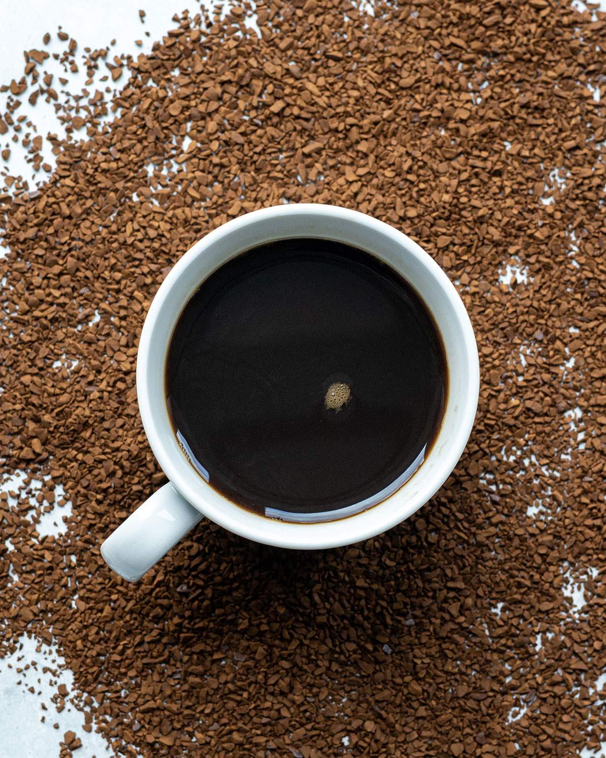 Overhead cup of black coffee surrounded by loose instant coffee granules on a light surface.