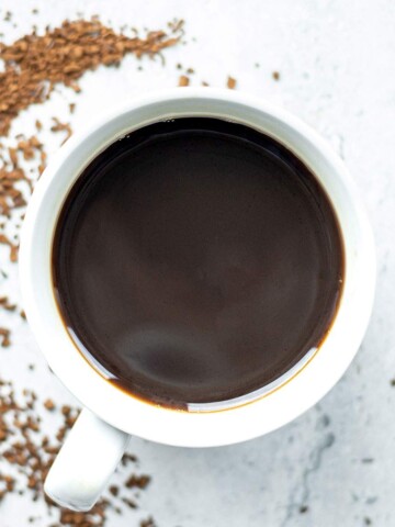 Overhead cup of black coffee beside a curved trail of instant coffee granules on a light surface.