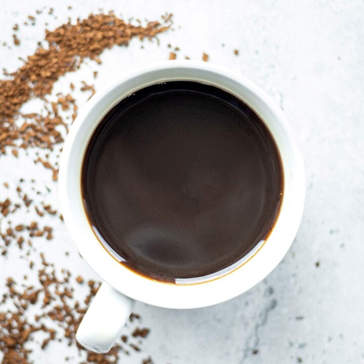 Overhead cup of black coffee beside a curved trail of instant coffee granules on a light surface.