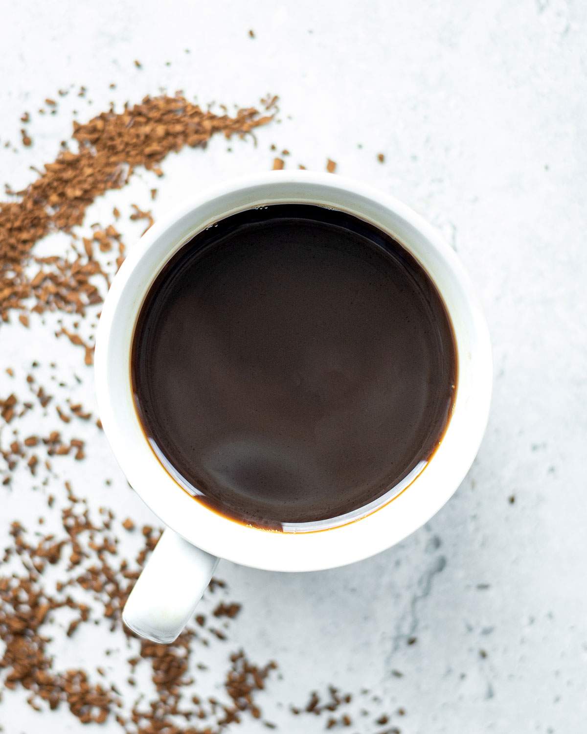 Overhead cup of black coffee beside a curved trail of instant coffee granules on a light surface.